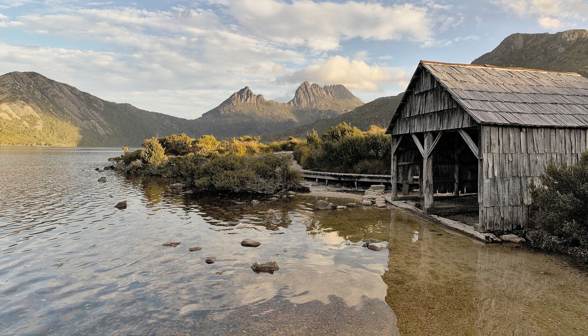 The jagged twin summits of Cradle Mountain above Dove Lake, with a weathered timber boatshed and low alpine scrub on the shore in evening light, Tasmania