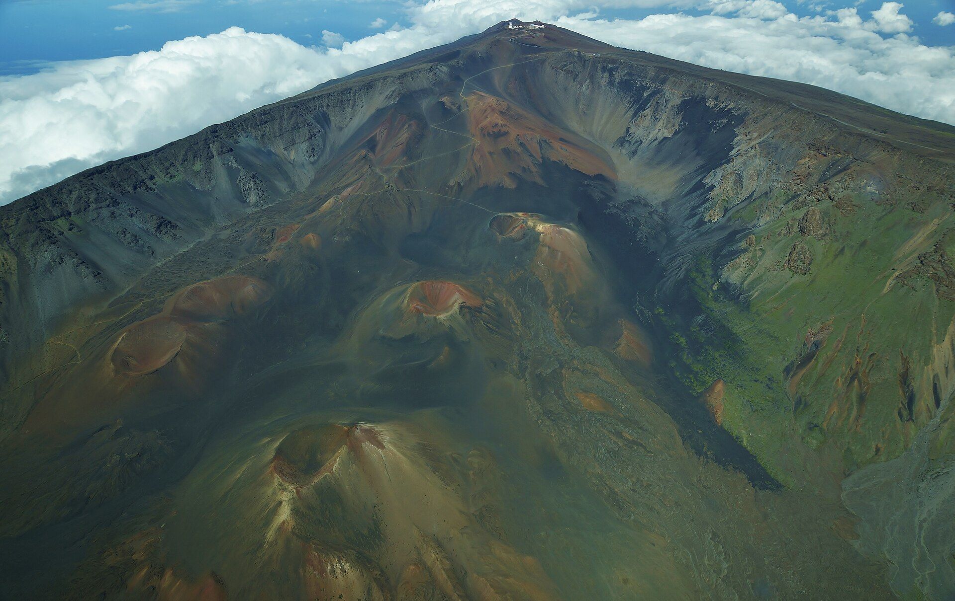 Aerial view down onto the summit depression of Haleakalā, with rust-red and grey cinder cones on the floor below steep walls and the summit road and observatories on the rim above a layer of white cloud