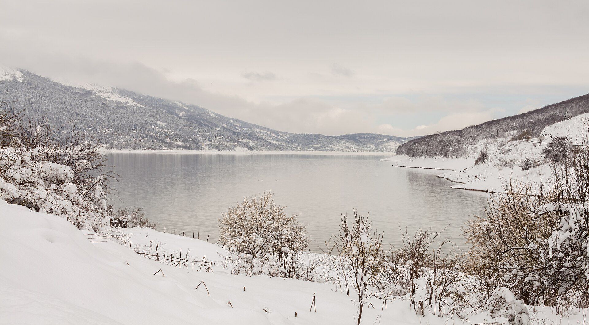 Lake Mavrovo in winter, its shores under snow and forested mountains rising into low cloud, Mavrovo National Park, North Macedonia