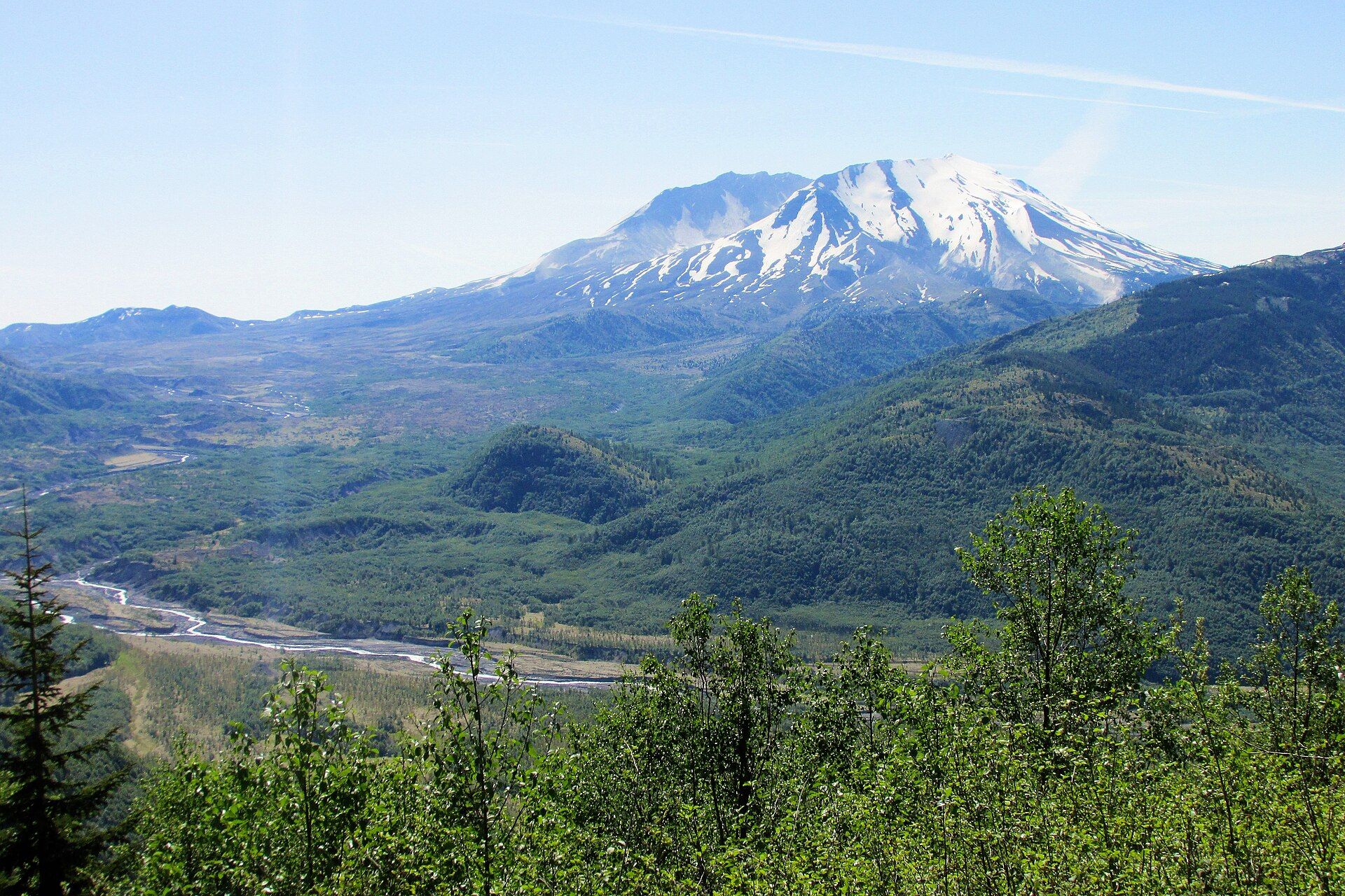 Snow-streaked Mount St. Helens with its collapsed north side open toward the viewer, above the braided channel of the Toutle River and green, regrowing forest in the valley below
