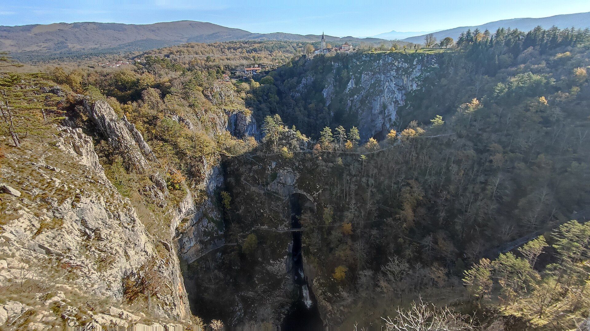 The deep, cliff-walled collapse dolines of Velika and Mala dolina with the Reka River falling into the cave mouth below and the village church of Škocjan on the rim, Slovenia