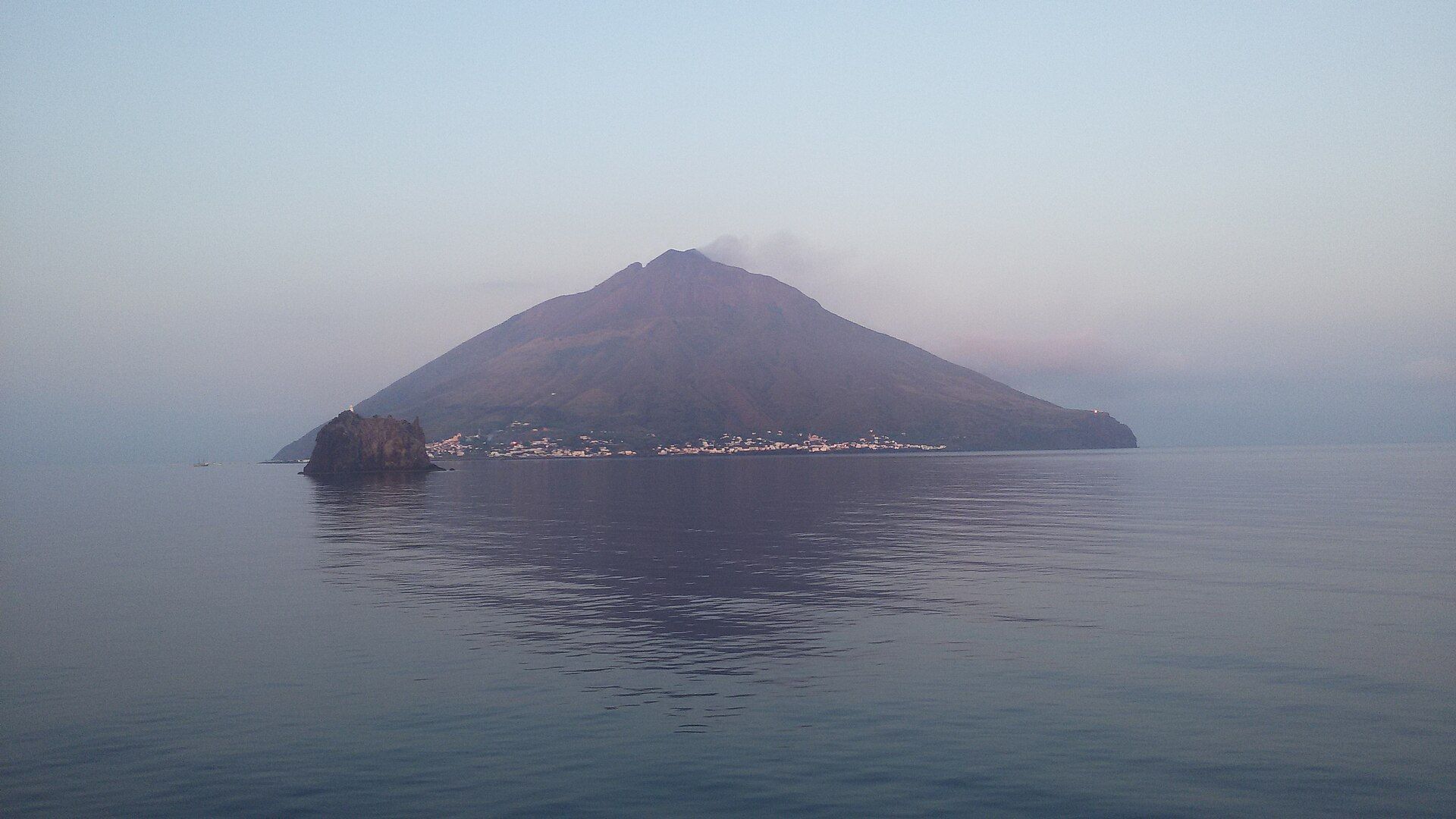 The cone of Stromboli rising from a calm, pale sea at dawn, a wisp of volcanic gas drifting from its summit, the white houses of Stromboli village along the shore and the rock of Strombolicchio in front