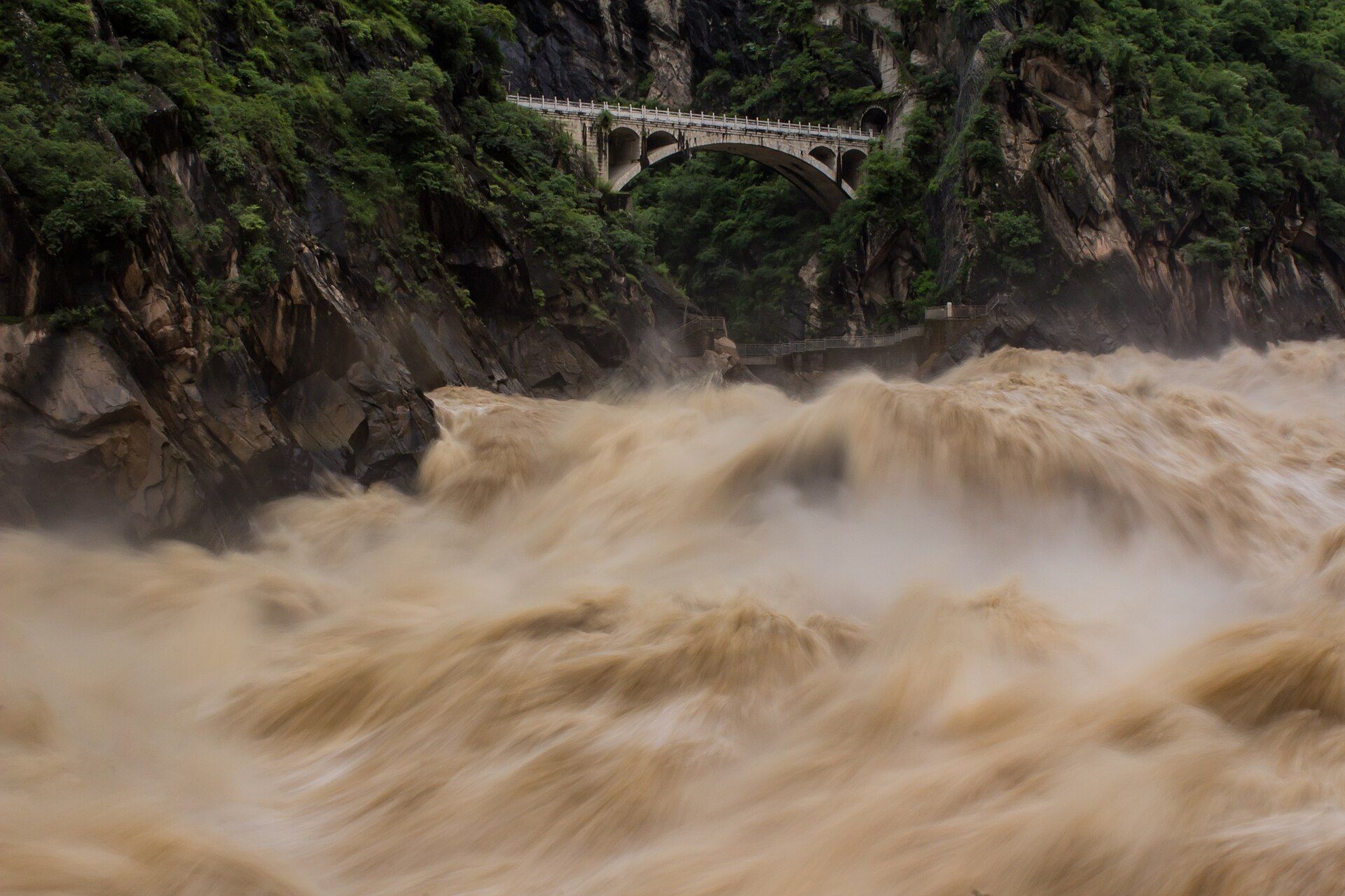 Brown floodwater of the Jinsha River surging through Tiger Leaping Gorge beneath a stone bridge, Yunnan, China
