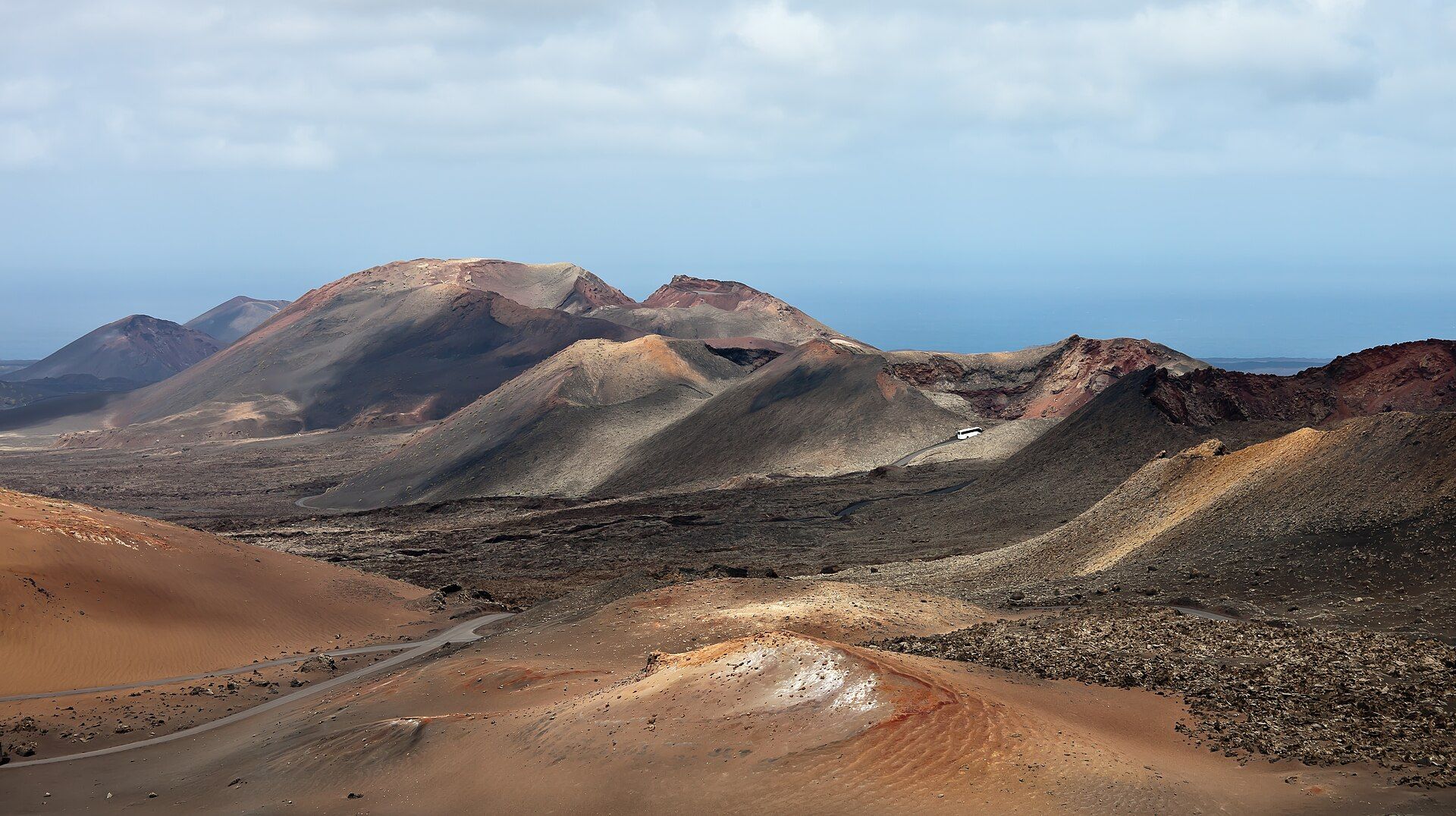 Rust-red, ochre and grey volcanic cones rising above a dark lava field, with a white coach on the winding Ruta de los Volcanes road and the Atlantic on the horizon, Timanfaya National Park, Lanzarote