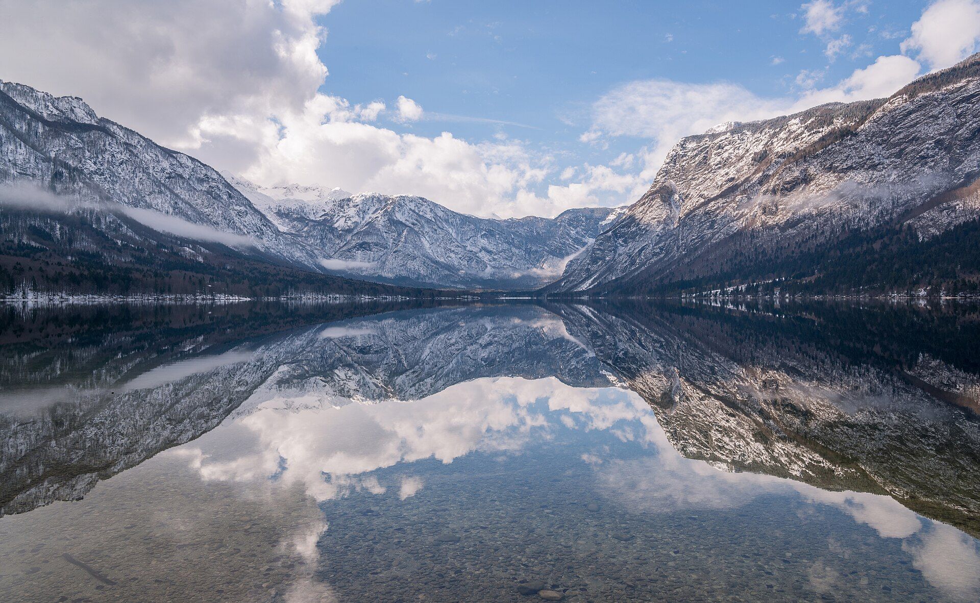 Snow-dusted limestone mountains of the Julian Alps mirrored in the still, clear water of Lake Bohinj under a cloudy sky, Triglav National Park, Slovenia